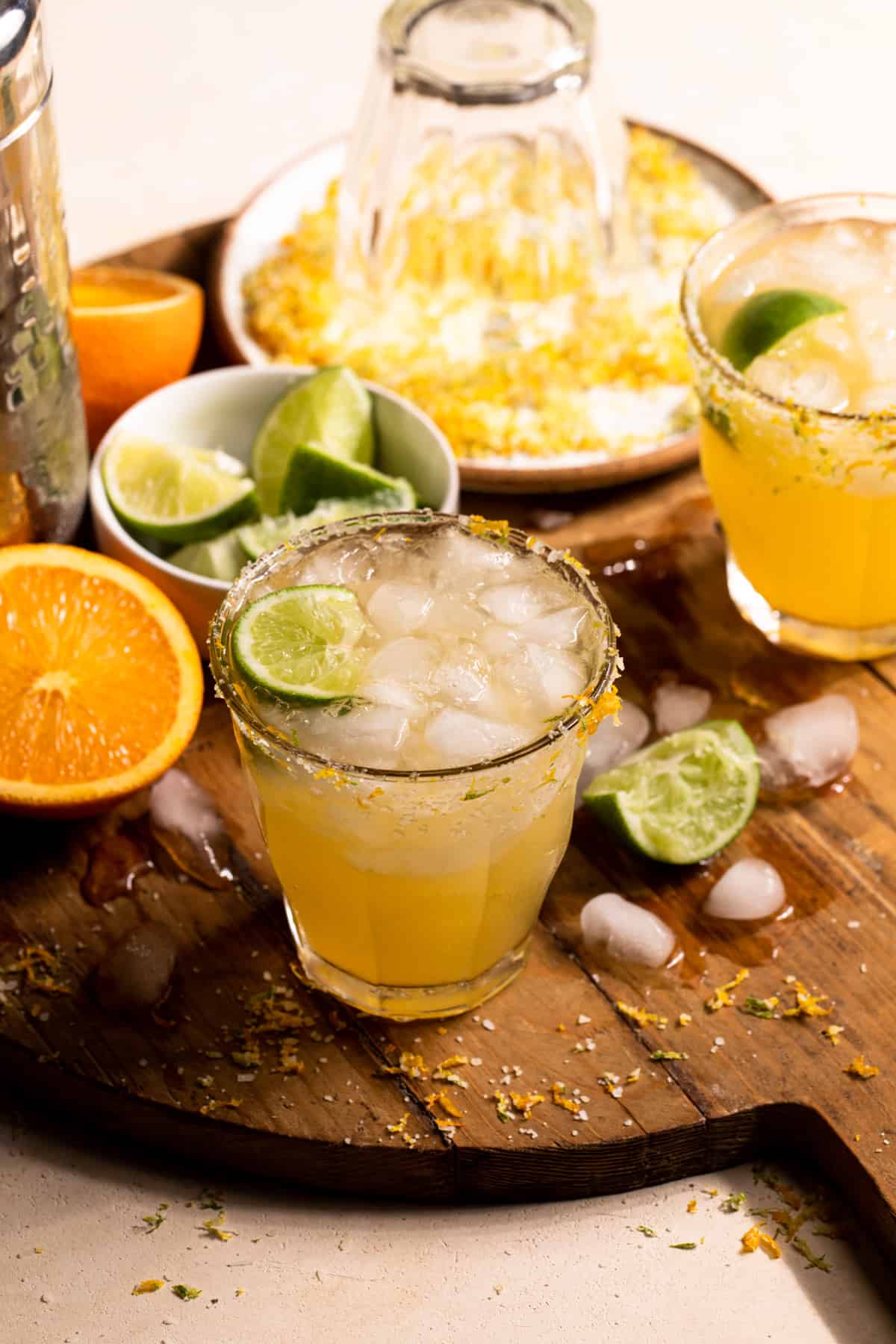 Texas orange and lime margaritas on a wooden board with ice and lime wedges around them.