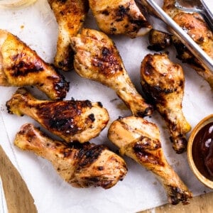 Grilled chicken legs on a wood cutting board with a bowl of bbq sauce and a beer.