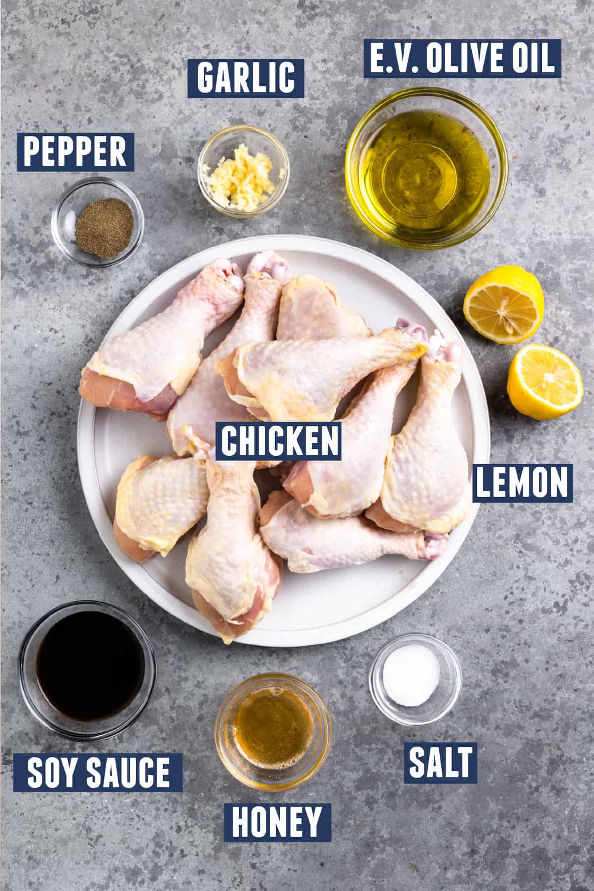 Ingredients needed to make grilled chicken legs laid on the counter, a plate of drumsticks, bowls of oil, garlic, salt and pepper, honey, soy sauce, and a lemon cut in half.