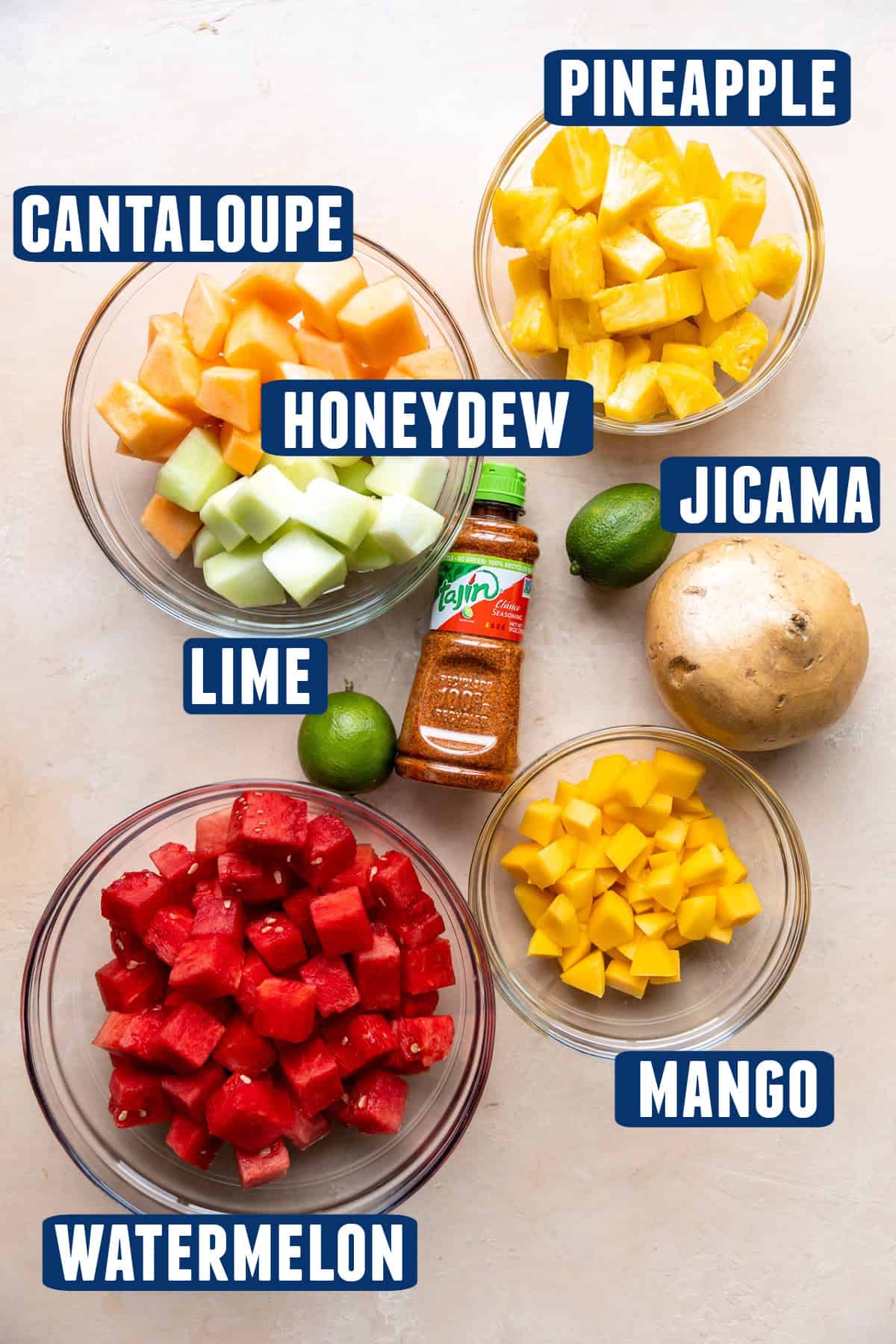 Ingredients needed to make a mexican fruit salad laid on the counter, bowls of chopped watermelon, mango, honeydew, cantaloupe, pineapple, a whole jicama, limes and bottle of Tajin.