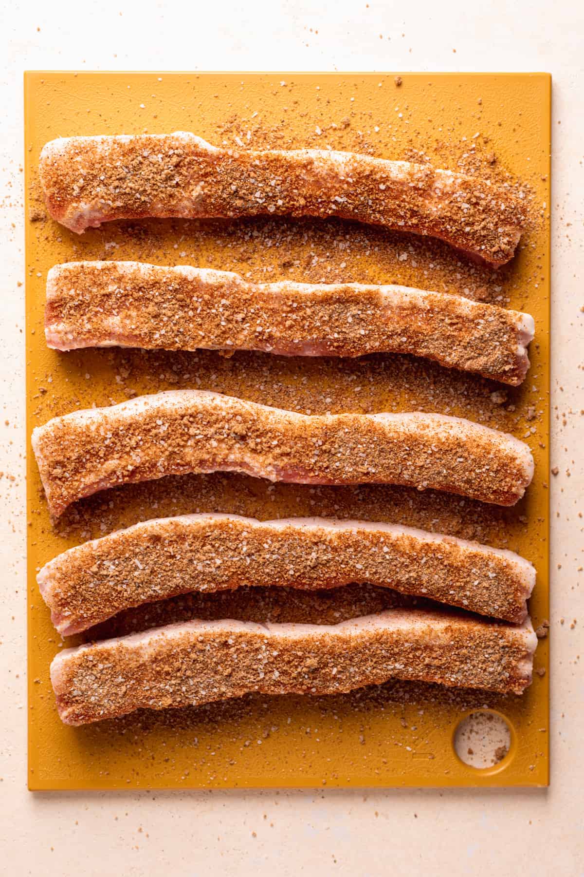 Strips of thick pork belly on a yellow cutting board sprinkled with pork dry rub.