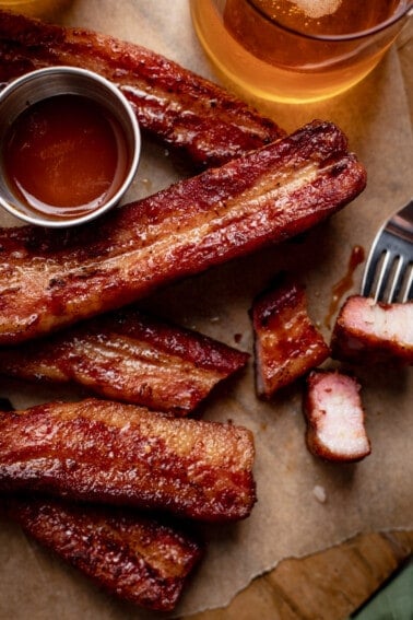Strips of smoked pork belly on brown parchment paper with a small bowl of BBQ sauce and a beer.