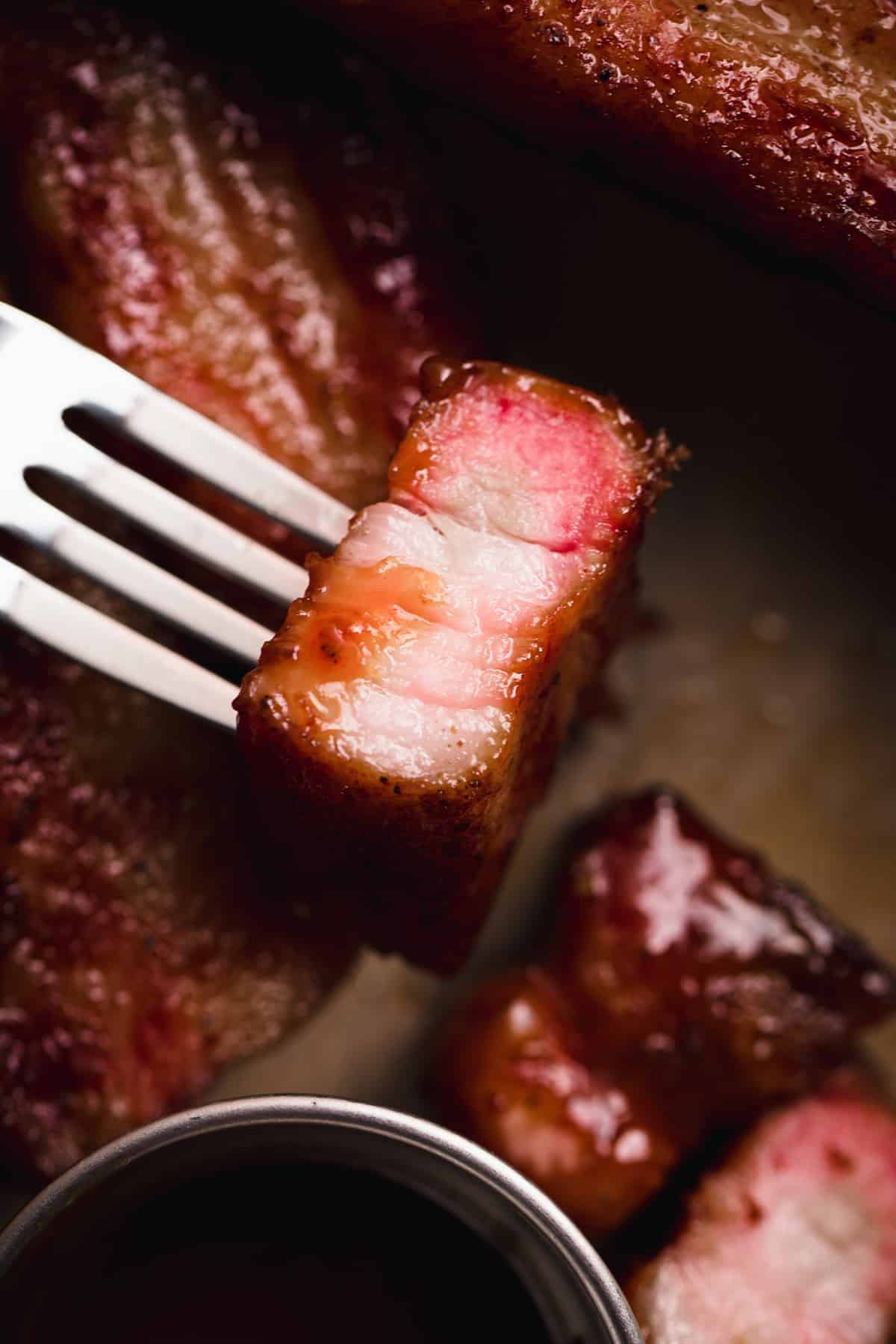 Chunk of thick pork belly sliced and on a fork showing a pink smoke ring and drizzled with some bbq sauce.