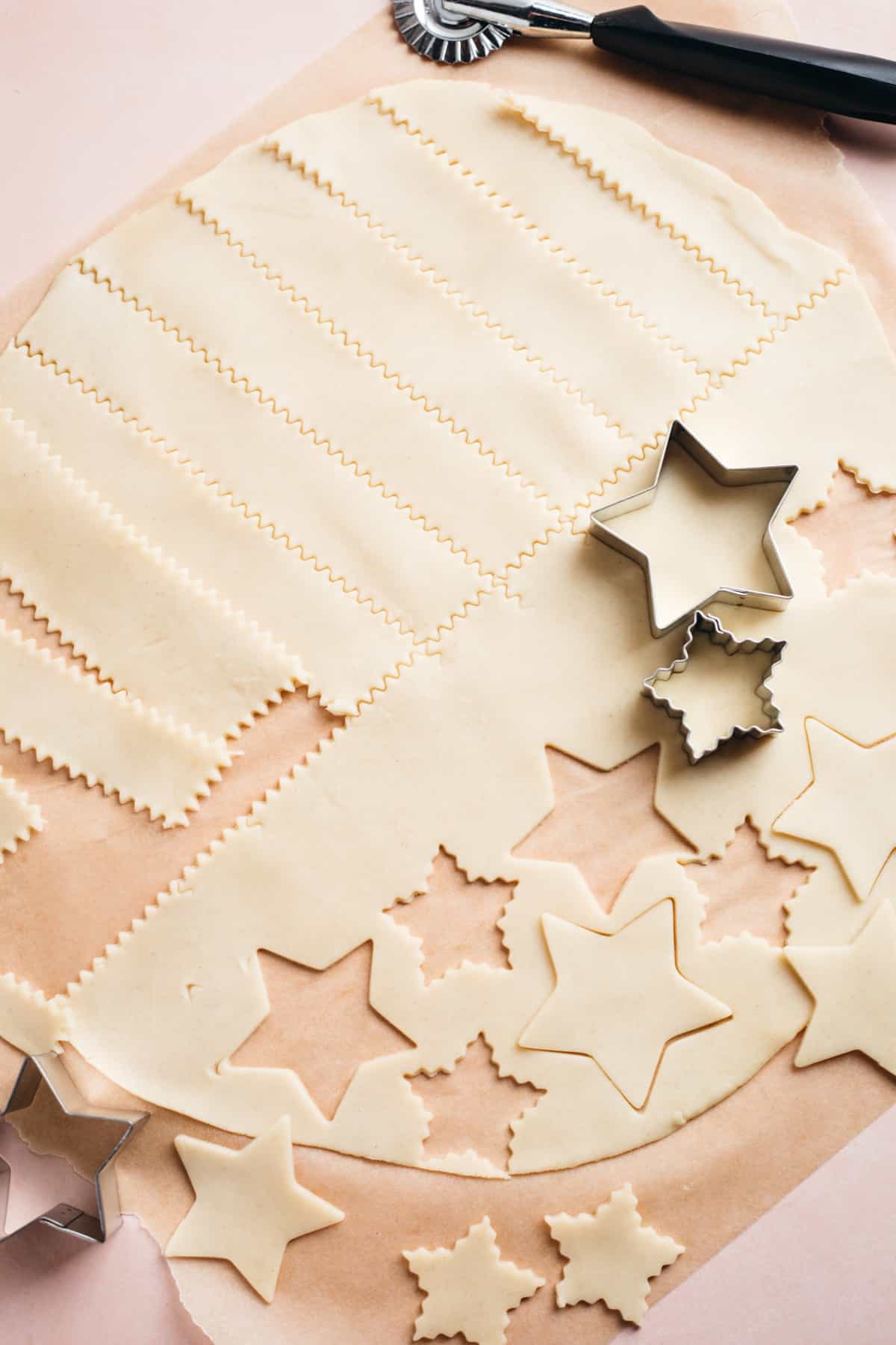 Pie crust rolled out on parchment paper and star cookie cutters and stripes being cut out.