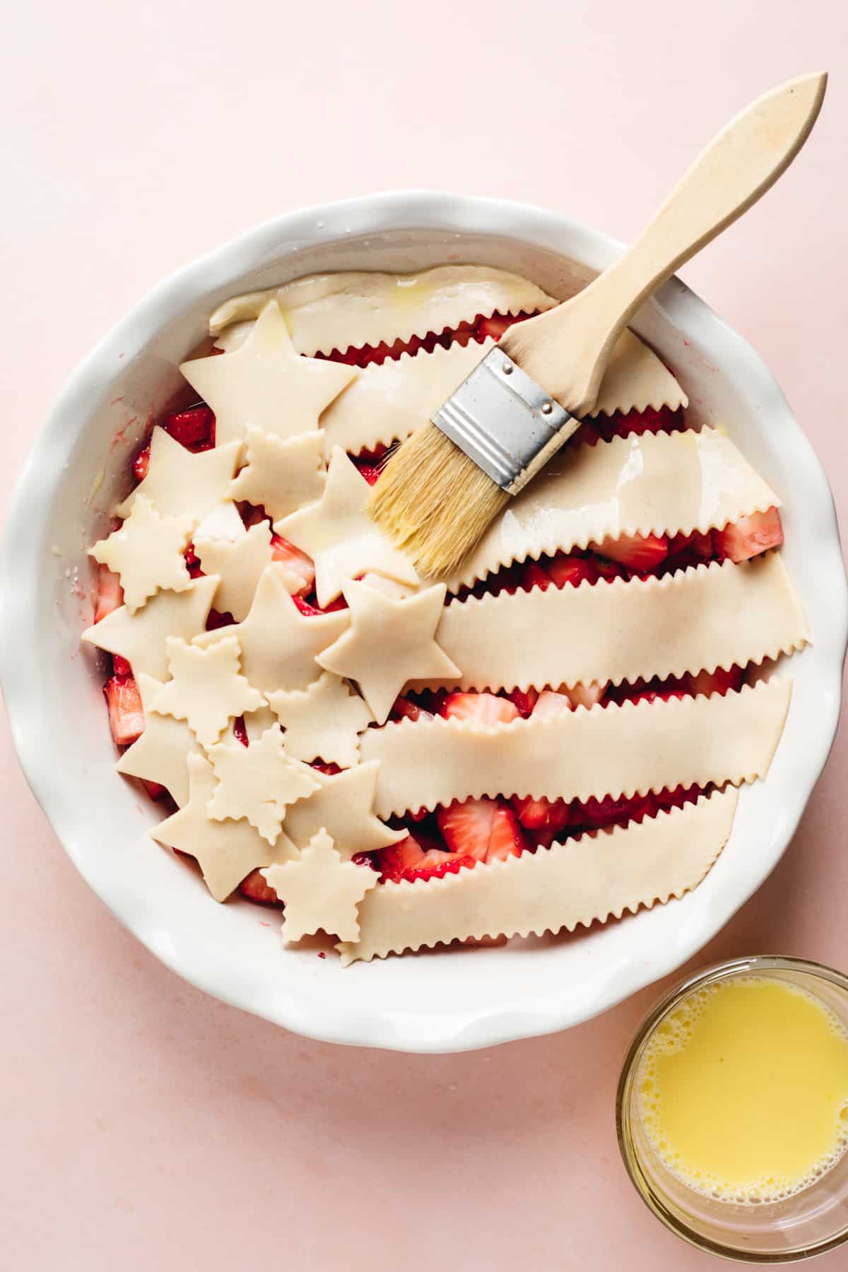 Pie dish filled with strawberry cobbler filling, topped with stars and stripes cut out of pie crust and a brush to put egg wash on. 