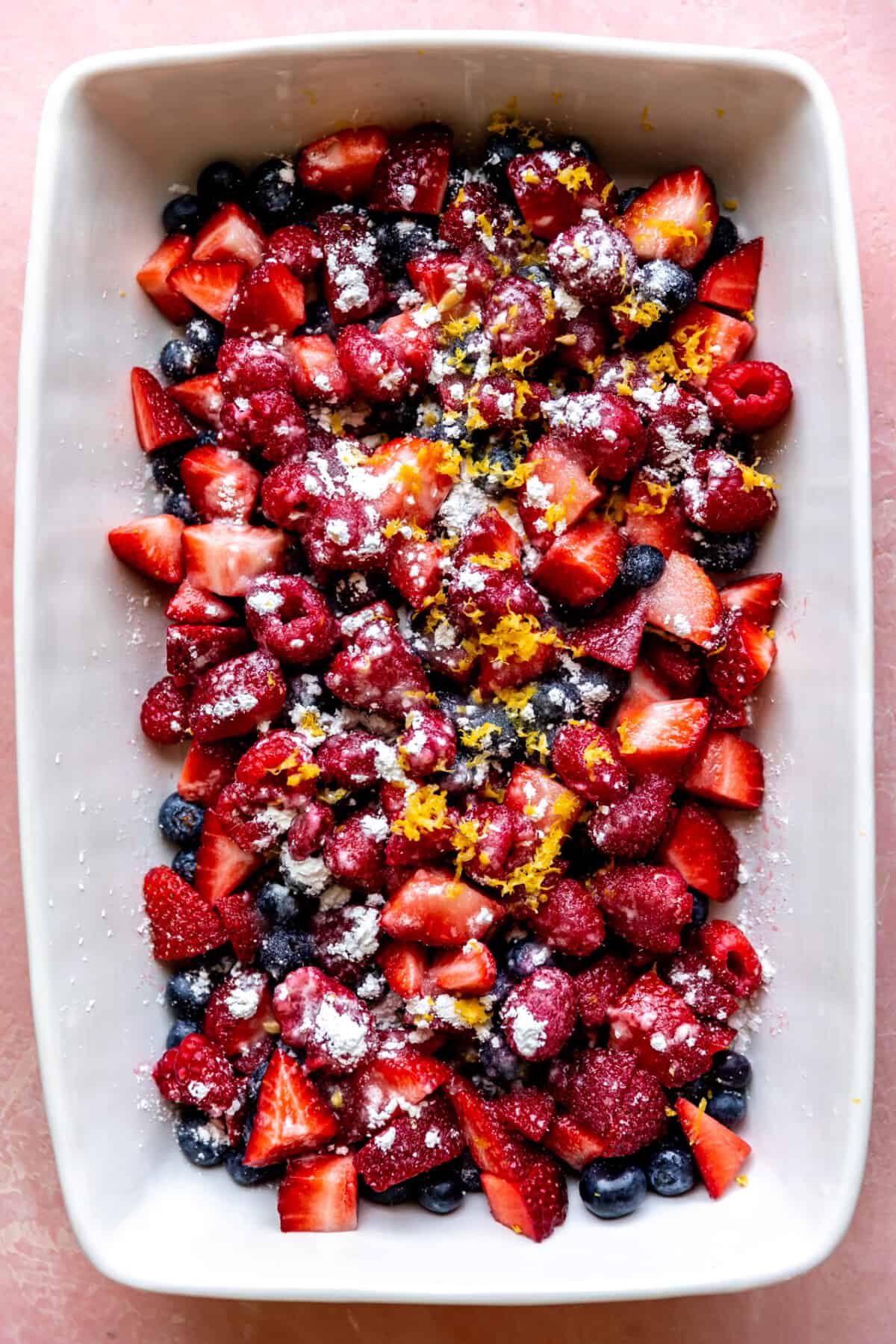 White baking dish filled with mixed berries topped with sugar, cornstarch and lemon zest.
