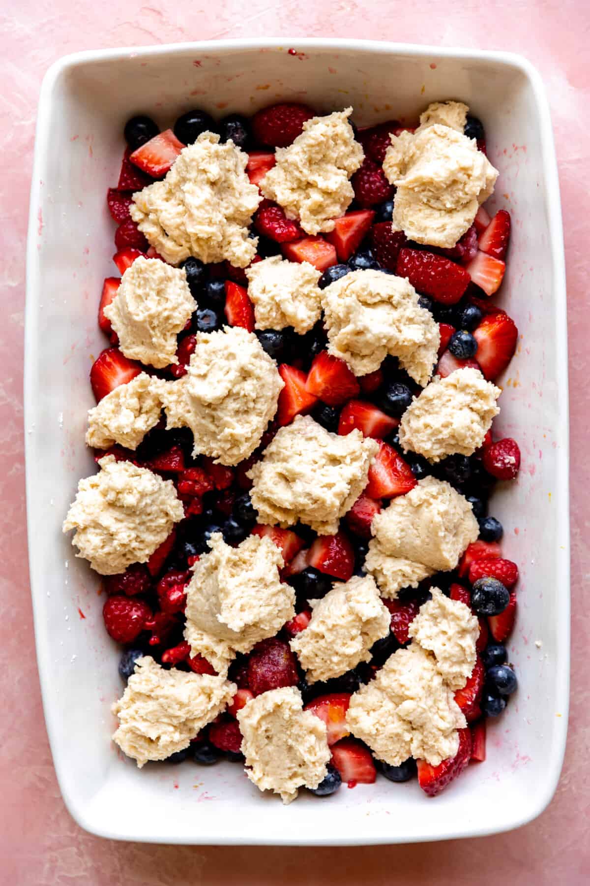 Cobbler batter spooned on top of mixed berries in a white baking dish. 