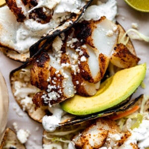 Grilled fish tacos on charred corn tortillas served with avocado slices and cilantro crema drizzled on top.