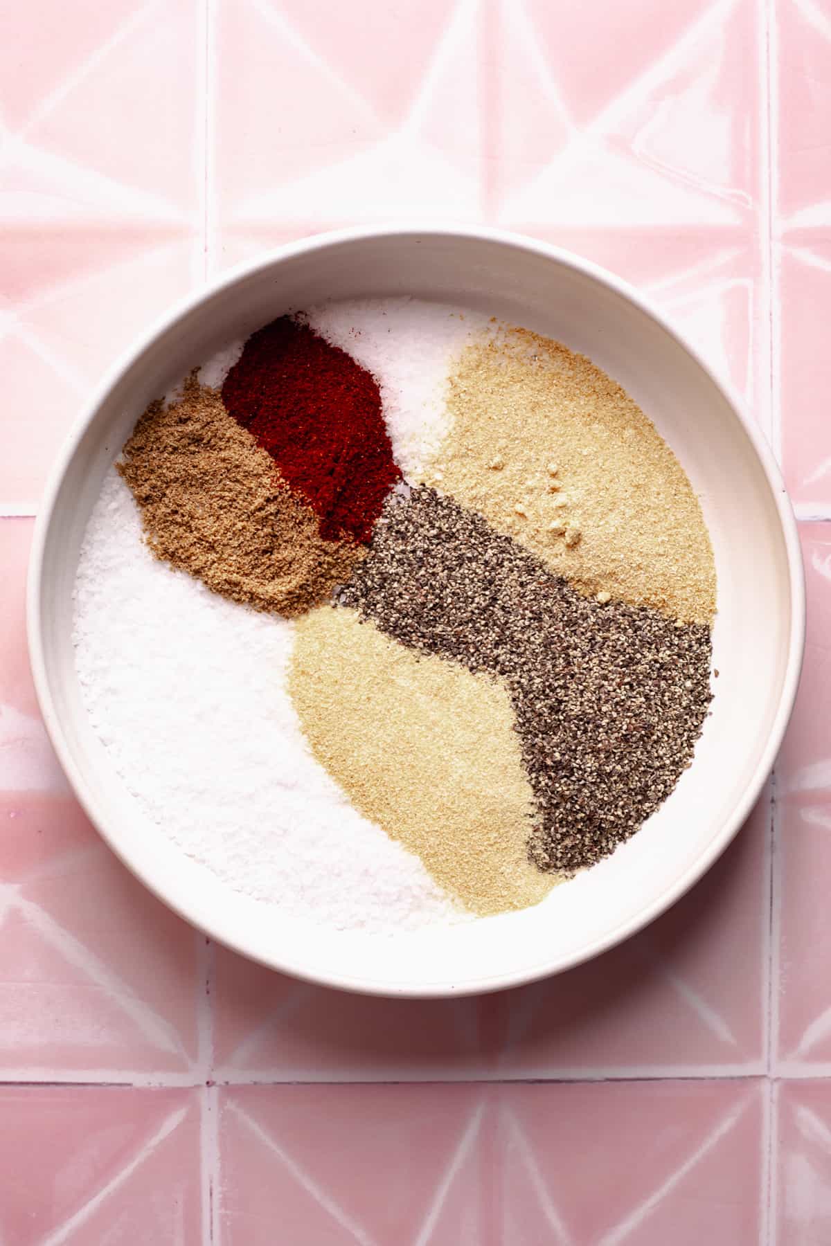 Spices in a white bowl on pink tile.
