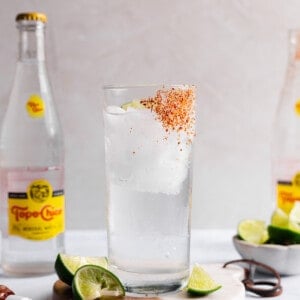 A glass cup with tajin on the side and ice with clear liquid and a lime wedge inside.