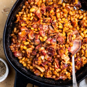 Cast iron skillet filled with baked beans topped with crispy bacon and coarse ground black pepper.