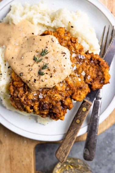 Chicken fried steak on a white plate with mashed potatoes and cream gravy on top.