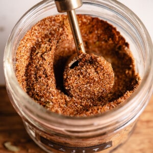 Jar filled with red spice mix with a wooden handled teaspoon scooping it up.