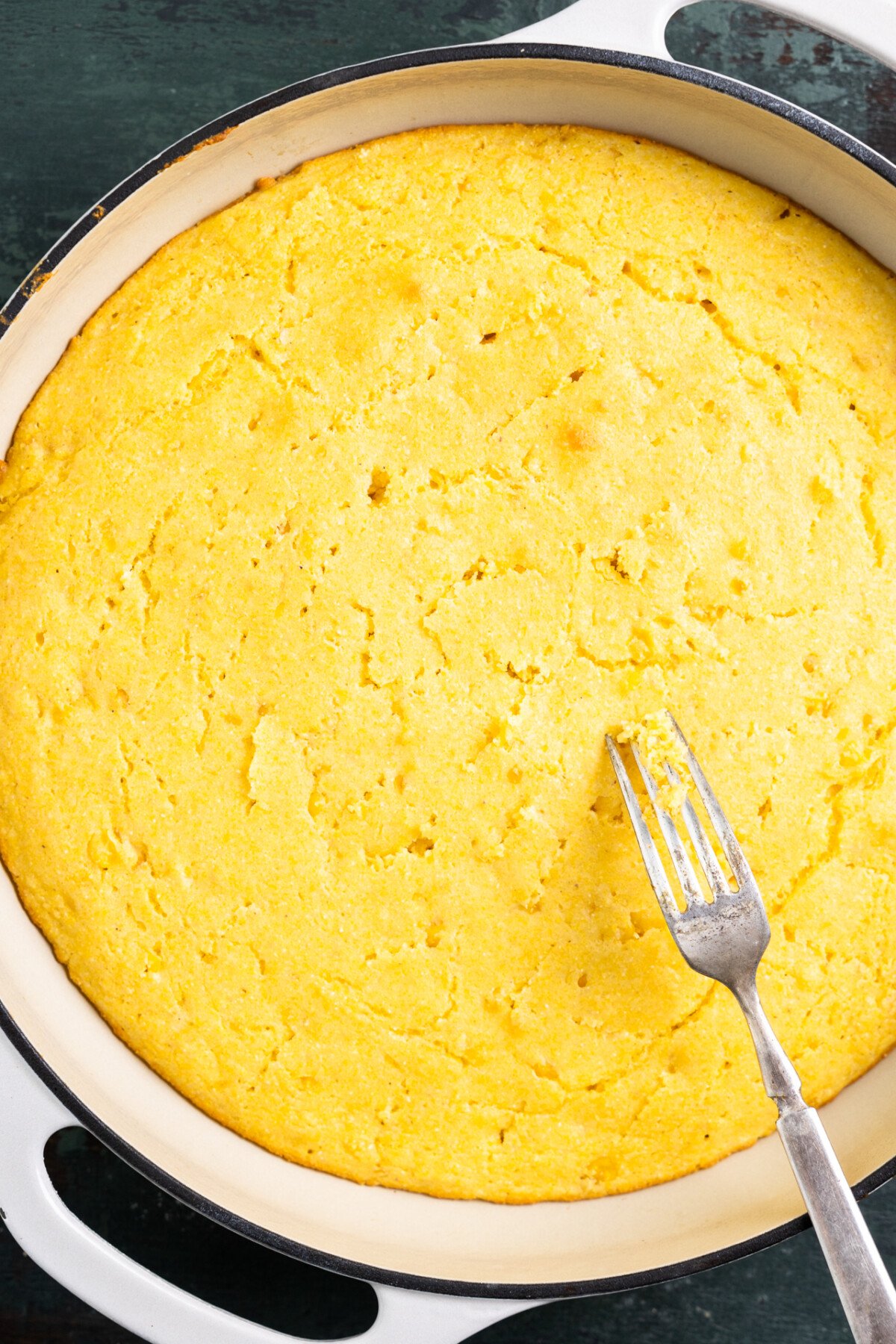 Baked cornbread in a white baking dish with a fork poking holes in the top. 