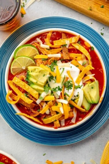 Bowl filled with chicken tortilla soup, topped with crispy tortilla strips, avocado, sour cream and slices of lime.