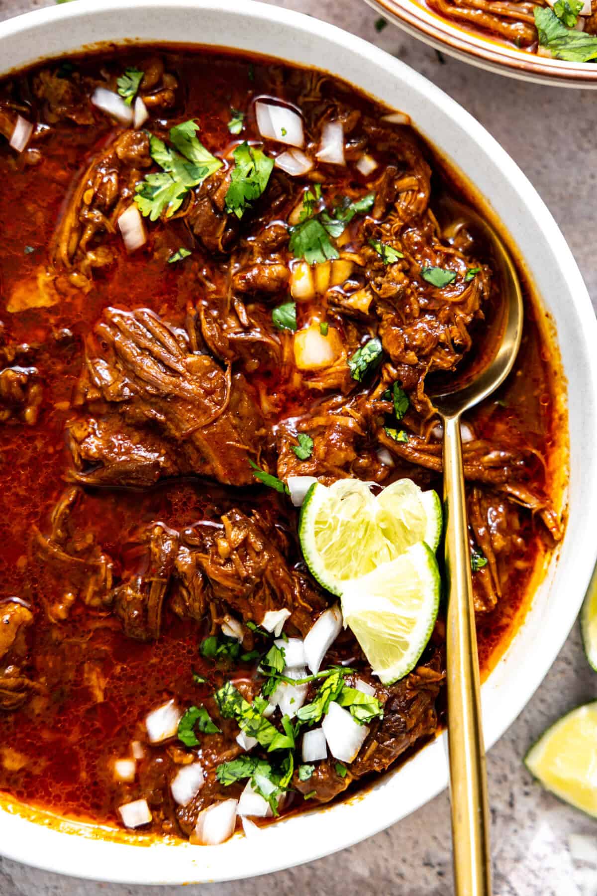 Bowl of birria served with diced onion and cilantro, topped with lime wedges. 
