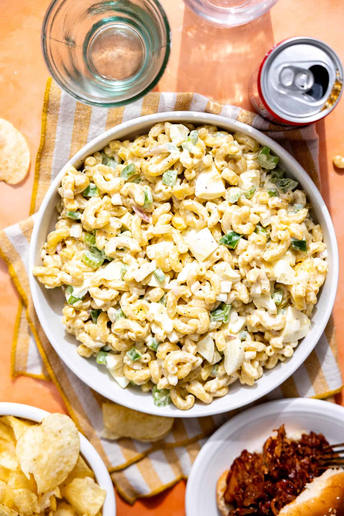 Bowl of macaroni salad on a picnic table with chips and pulled pork nearby.