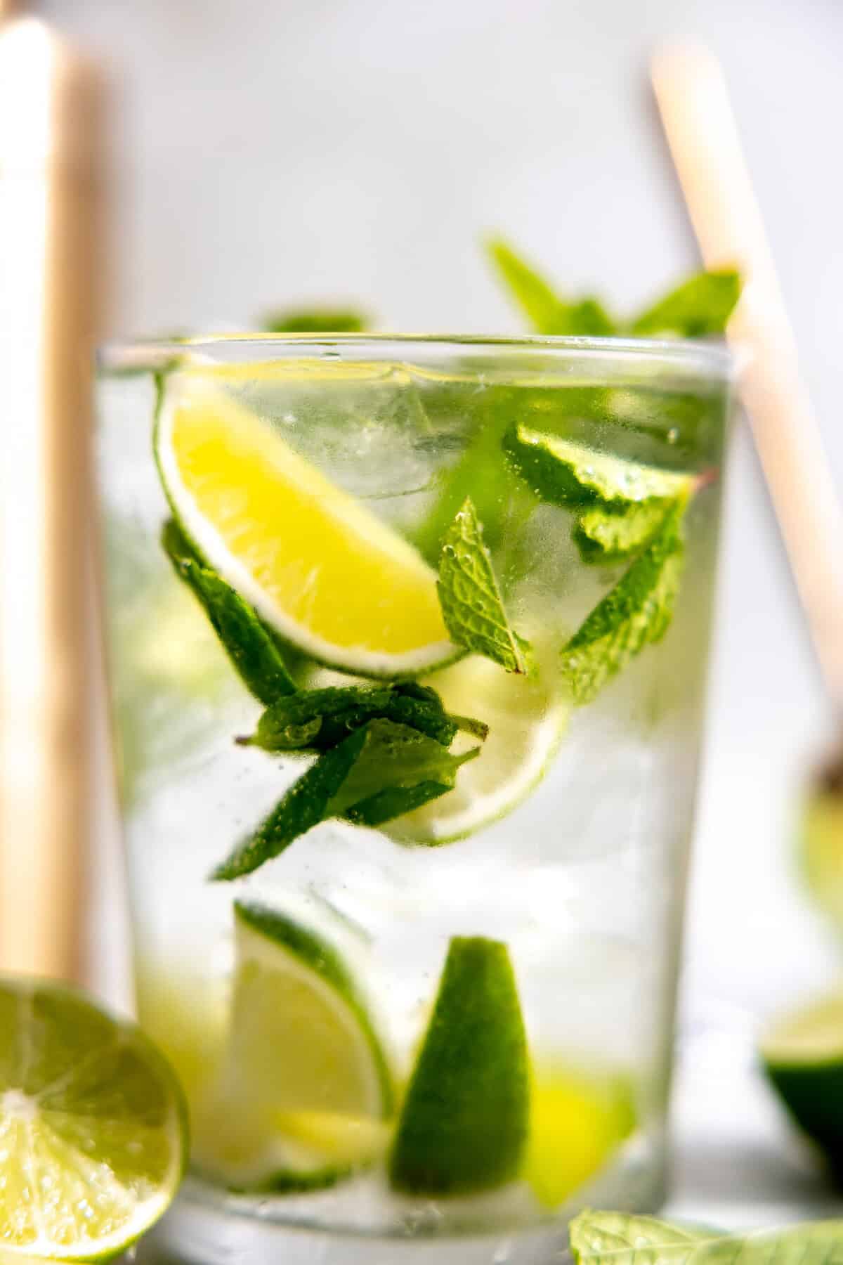 Up close of a glass filled with ice, mint leaves, lime wedges, and mojito cocktail. 