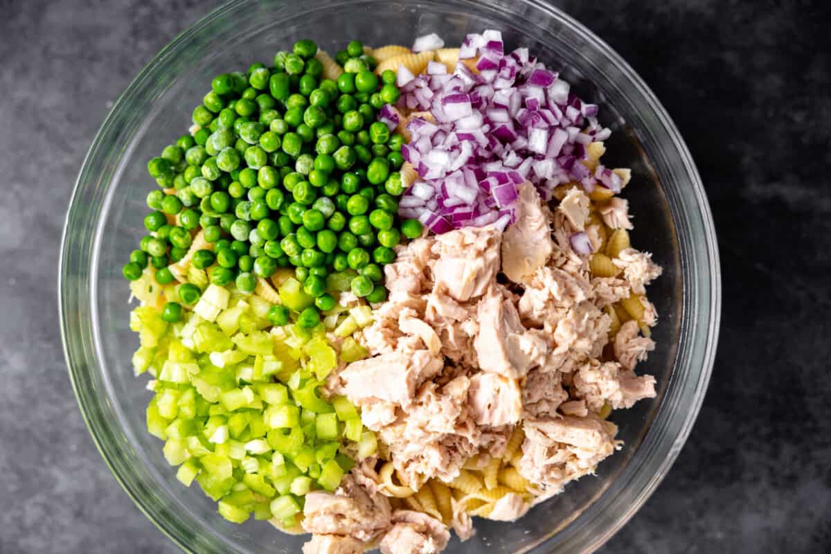 Cooked pasta, tuna, red onion, peas, celery in a glass bowl before mixing.