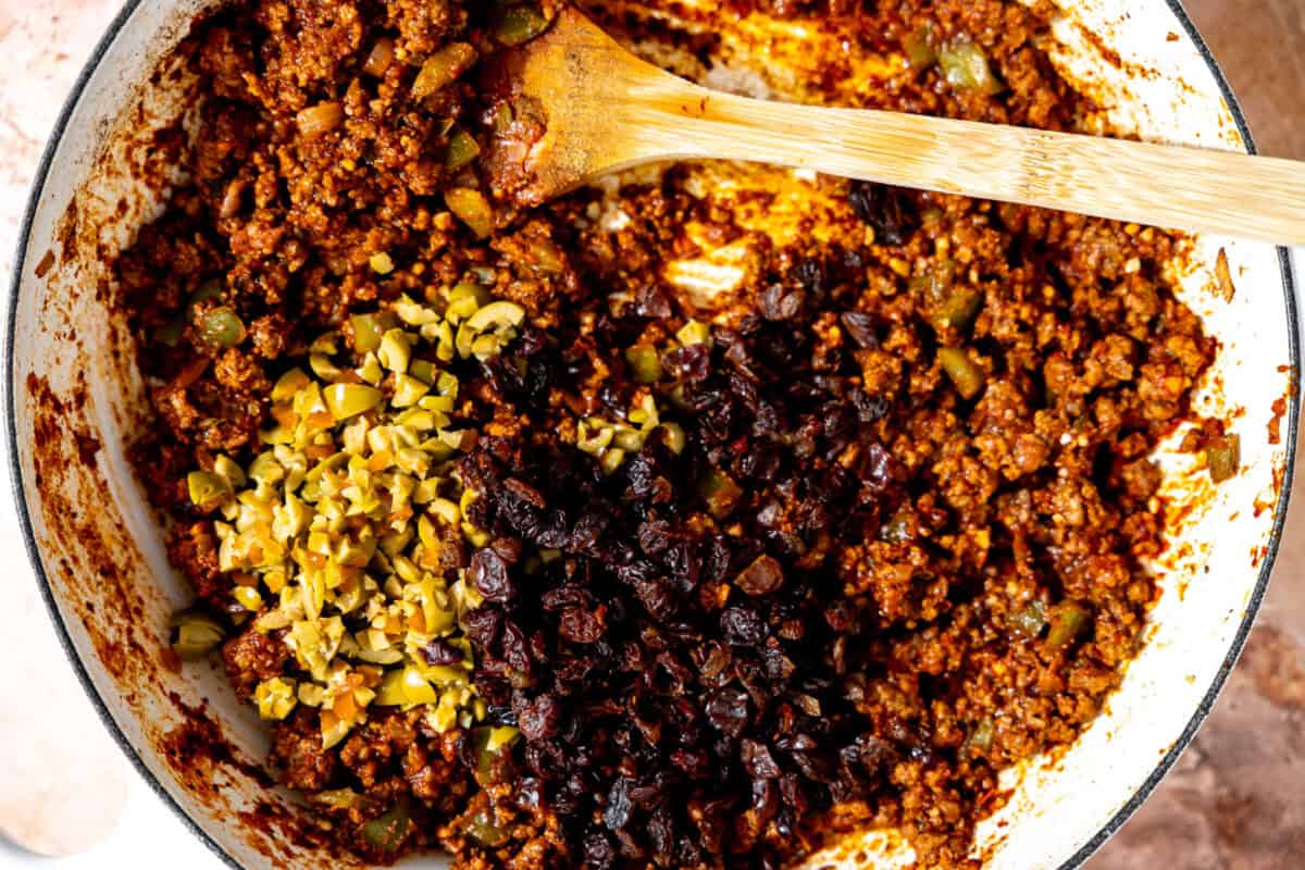 Adding raisins and green olives to a skillet of picadillo. 