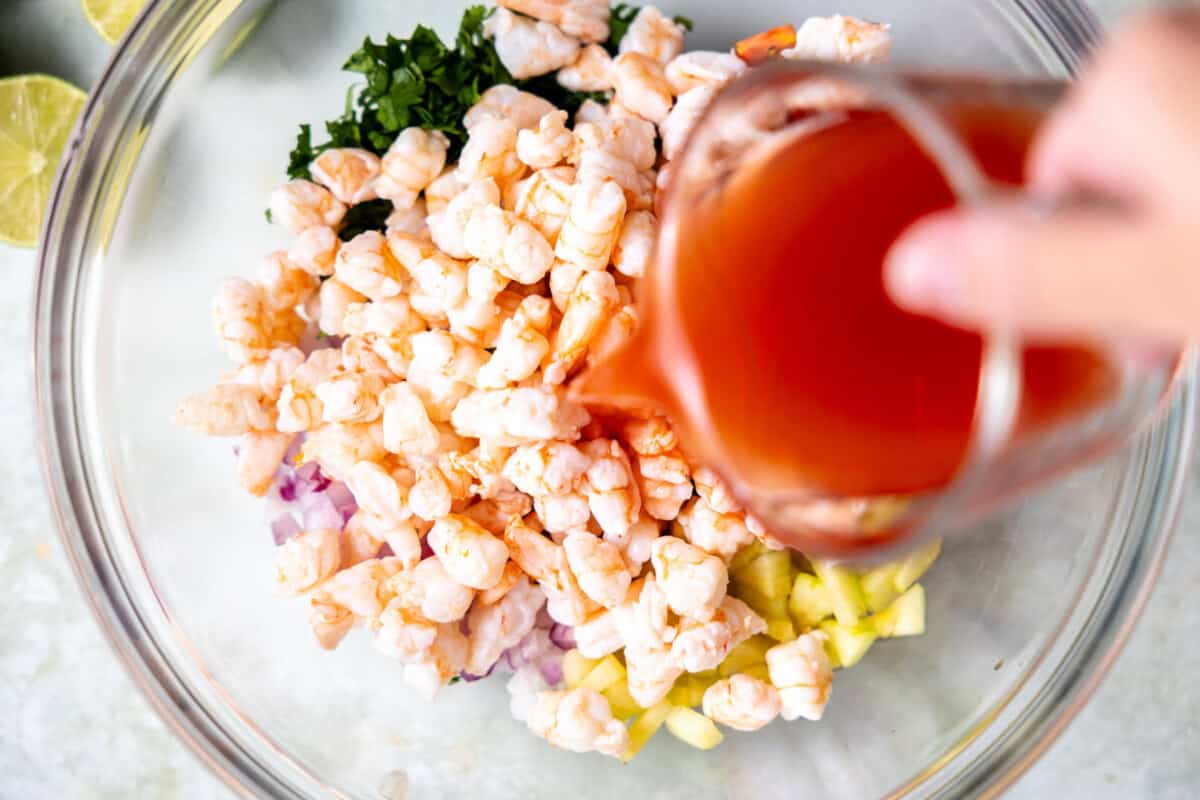 Pouring Clamato juice over shrimp and veggies in a bowl to make ceviche. 