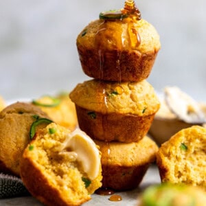 Golden jalapeño cornbread muffins topped with melted cheddar, featuring bits of jalapeño for a spicy kick, arranged on top of each other and being drizzled with honey.