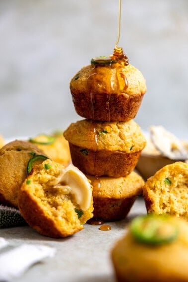 Golden jalapeño cornbread muffins topped with melted cheddar, featuring bits of jalapeño for a spicy kick, arranged on top of each other and being drizzled with honey.