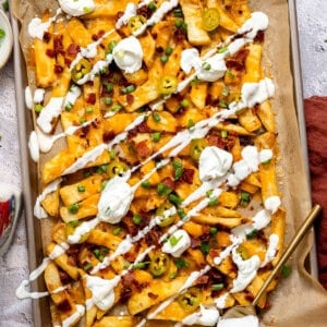 Golden, crispy French fries loaded with melted cheese, bacon, green onions, and a drizzle of sour cream on a baking tray.