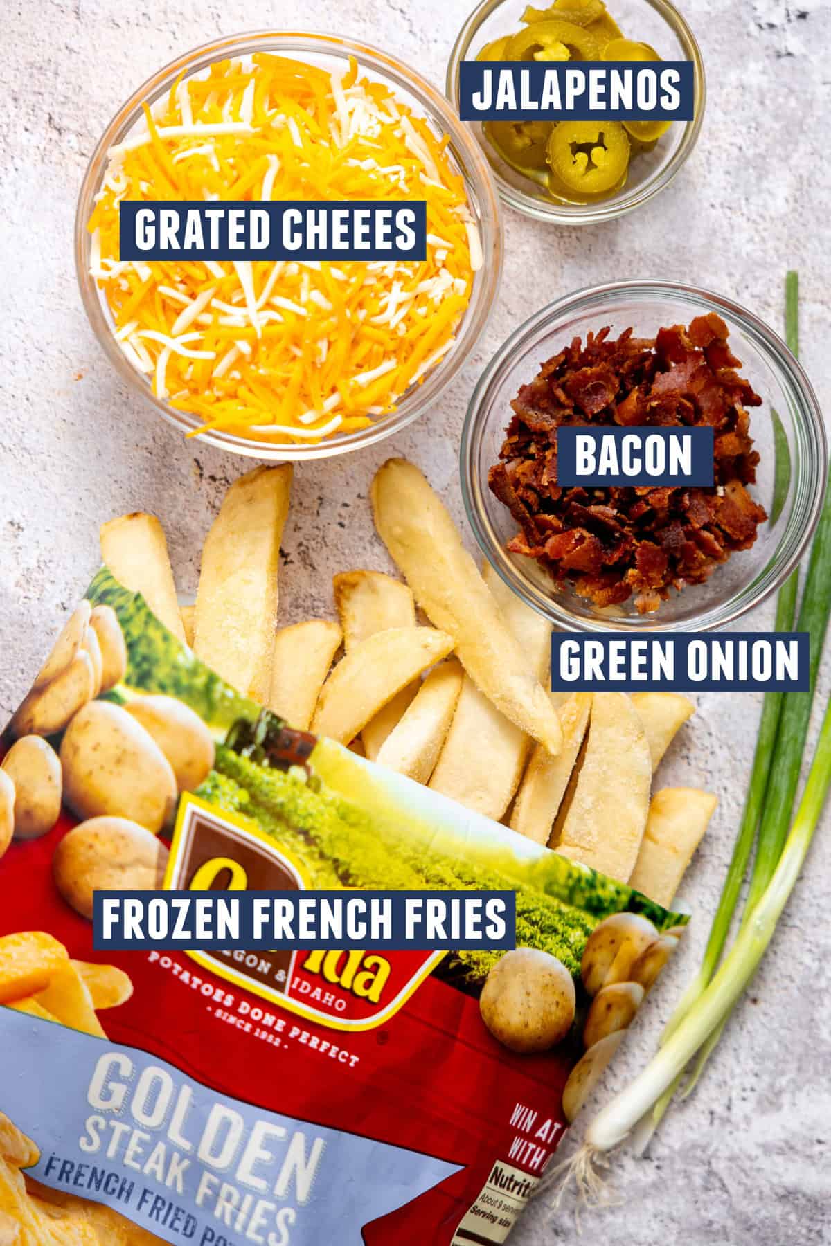 Ingredients laid out for making loaded French fries including: frozen French fries, grated cheese, crumbled bacon, pickled jalapeños, and green onions. 