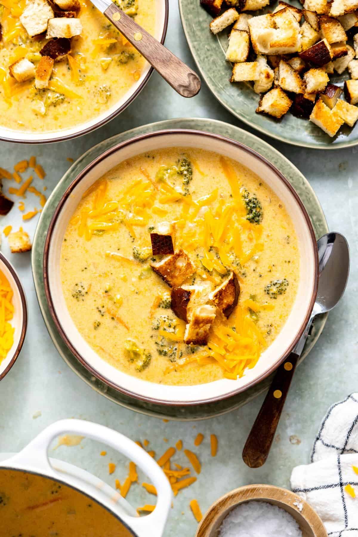 A creamy bowl of broccoli cheddar soup topped with shredded cheddar cheese and served with a side of crusty bread.