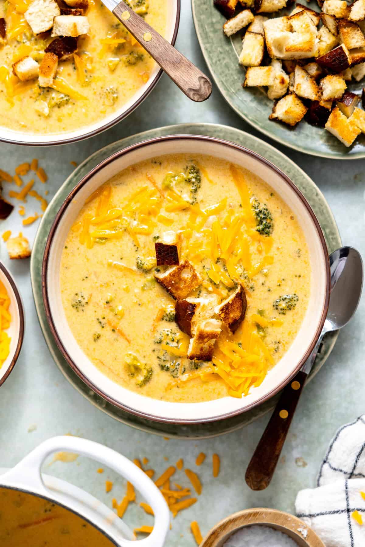 A creamy bowl of broccoli cheddar soup topped with shredded cheddar cheese and served with a side of crusty bread croutons. 