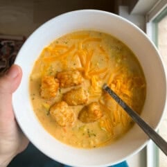 Bowl of broccoli cheddar soup topped with extra cheese and crispy tater tots. 