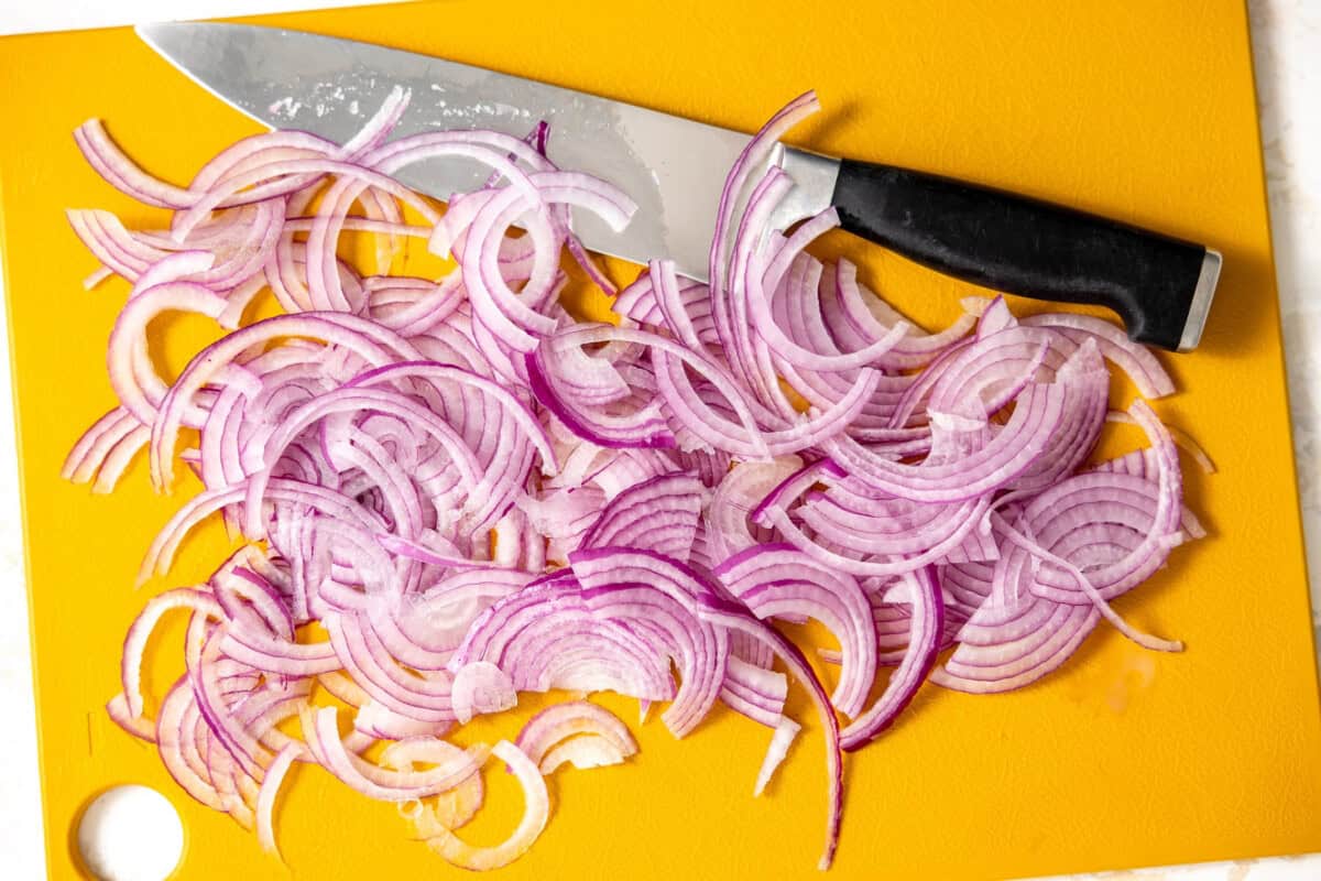 Red onion thinly sliced on a cutting board with a black-handled knife. 