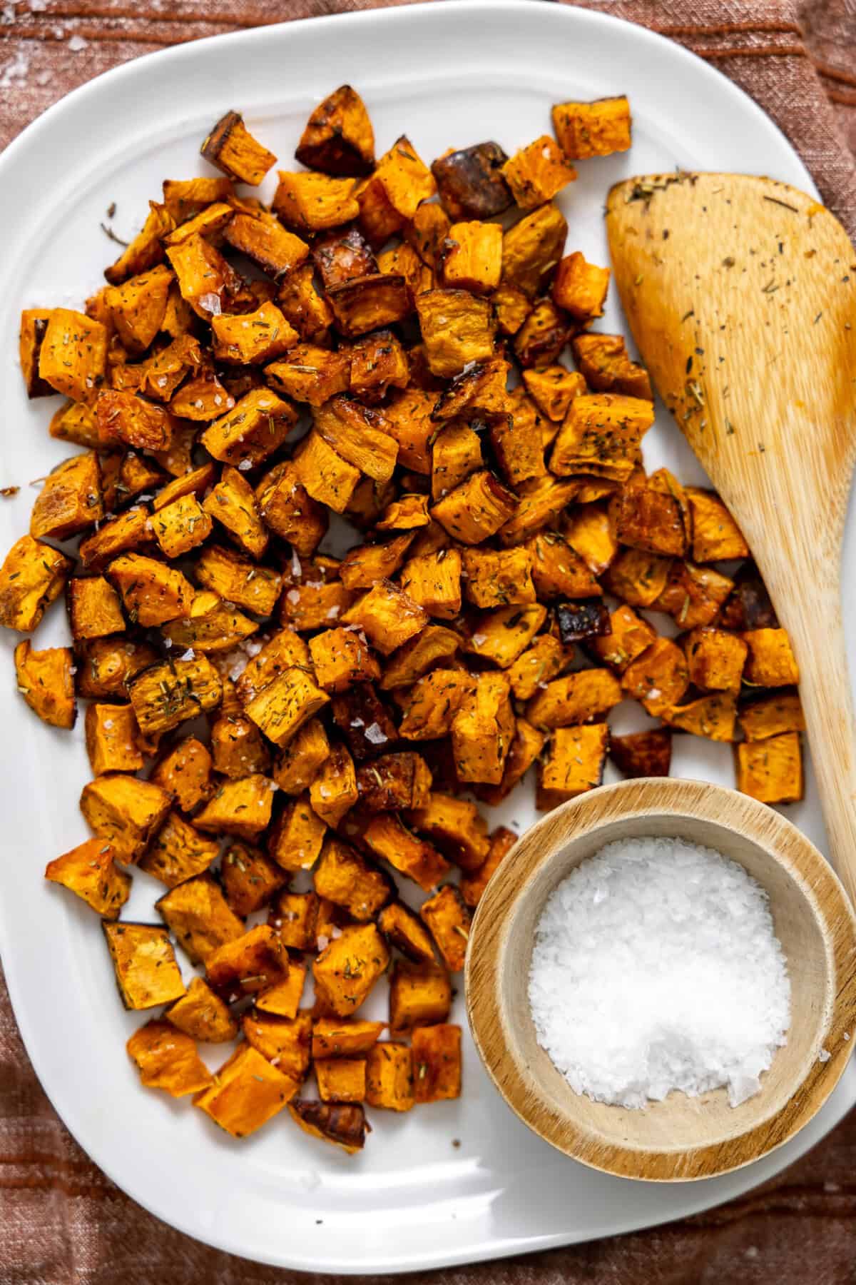 Plate of crispy roasted sweet potatoes with herbs and a small bowl of flaky salt with a wooden spatula. 