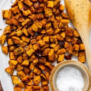 Plate of crispy roasted sweet potatoes with herbs and a small bowl of flaky salt with a wooden spatula.