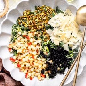 Kale and brussels sprout salad topped with apple, pistachios, dried cherries and parmesan in a white serving bowl.