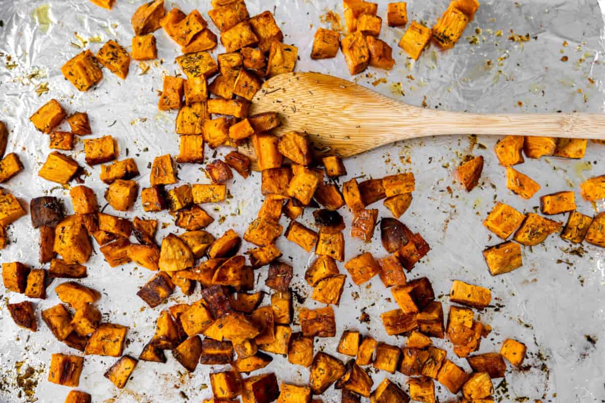 Roasted sweet potatoes on a baking sheet with a wooden spatula to stir around. 