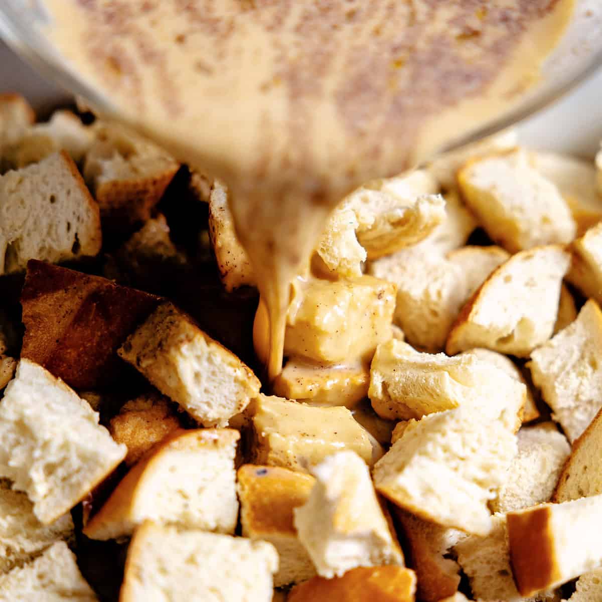 Pouring custard mixture over dried French bread cut into cubes in a baking dish. 