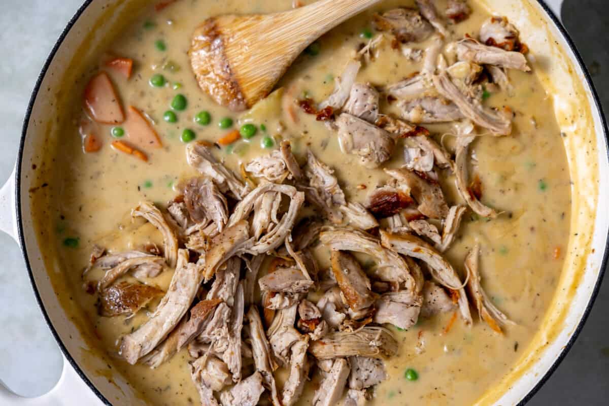 Shredded chicken being stirred into a creamy pot pie filling in a skillet, ready for baking