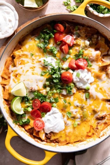 Ground beef enchilada skillet in a braising pan, topped with sour cream, fresh cilantro, diced onion, and lime wedges.