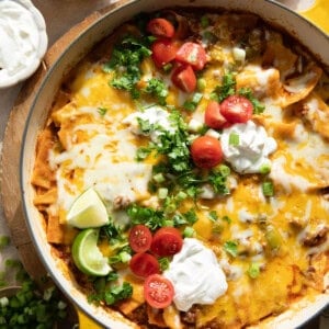 Ground beef enchilada skillet in a braising pan, topped with sliced tomatoes, fresh cilantro, sour cream, and lime wedges.