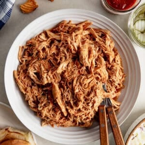 Shredded BBQ Chicken in a bowl with hamburger buns, pickles and sauce next to it.