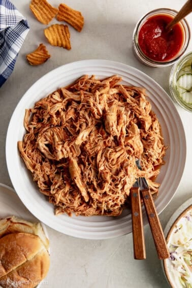 Shredded BBQ Chicken in a bowl with hamburger buns, pickles and sauce next to it.